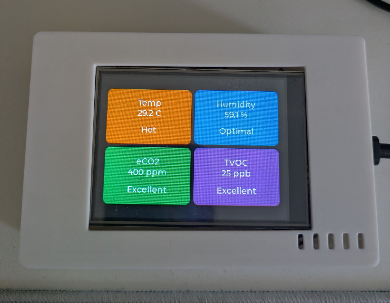 Top-down view of the monitor lying flat. The display shows a 2x2 grid of cards: orange Temp 29.2 C Hot, blue Humidity 59.1 % Optimal, green eCO2 400 ppm Excellent, purple TVOC 25 ppb Excellent. Five exhaust slots are cut into the lid below the screen.